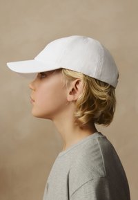 Casquette de baseball blanche en coton portée par une personne aux cheveux blonds courts, vêtue d'une chemise grise, vue de profil sur un fond neutre.