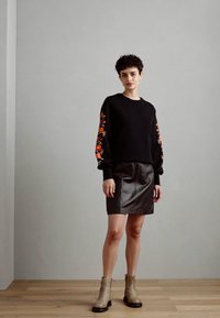 Suéter negro con bordado floral en las mangas, combinado con una falda de cuero negra. La modelo lleva botas de tobillo beige. Texturas suaves, corte holgado.