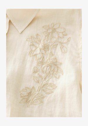 Cream-colored blouse featuring intricate beige floral embroidery along one side. Soft, lightweight fabric with a structured collar.