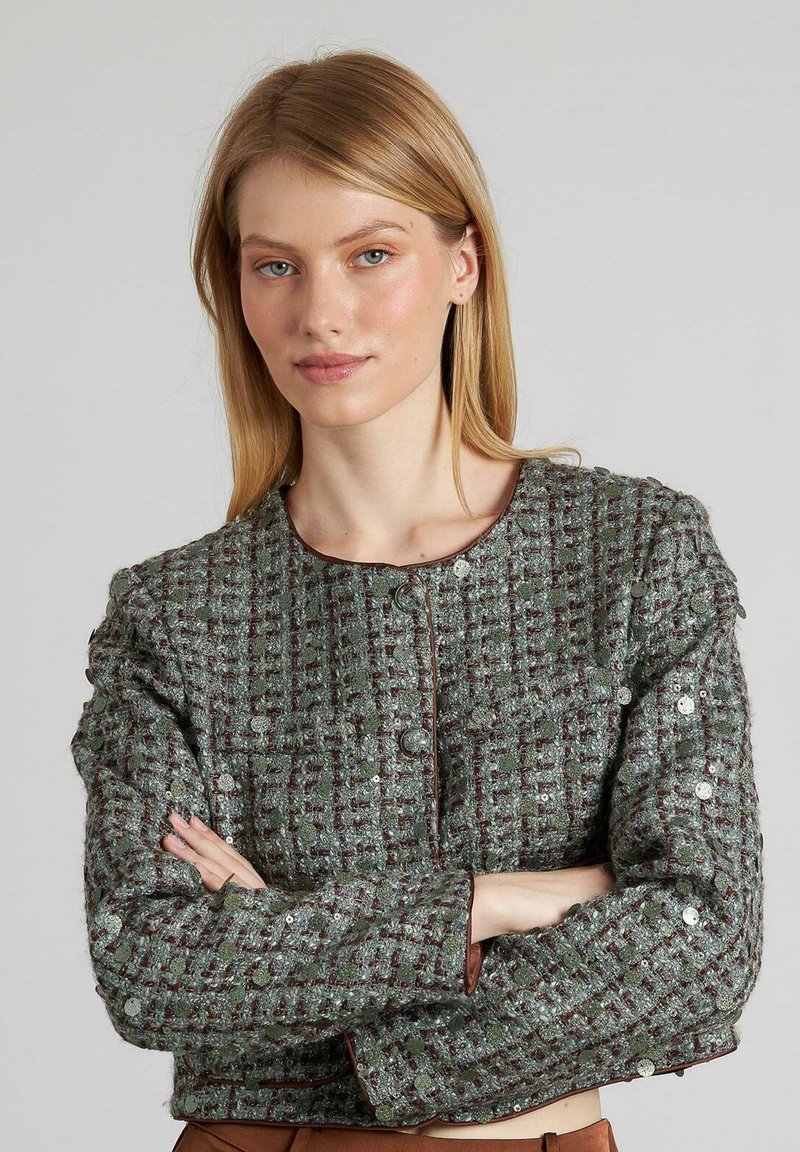 Grüne Tweedjacke mit einem strukturierten Muster, runder Halsöffnung, Frontknöpfen und metallischen Akzenten. Lange Ärmel mit leichtem Puffdetail.