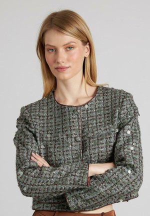 Chaqueta de tweed verde con un patrón texturizado, que cuenta con un cuello redondo, botones delanteros y acentos metálicos. Mangas largas con un leve detalle de volumen.