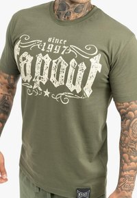 Olive zelené bavlnené tričko so zničeným bielym grafikou "Tapout" a logom, okrúhlym výstrihom a krátkymi rukávmi.