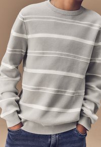 Pull en maille gris avec des rayures blanches horizontales, encolure ras du cou et poignets côtelés, coupe décontractée, associé à un jean en denim bleu.
