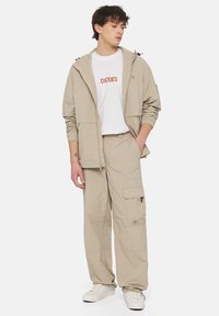 Beige lightweight jacket with drawstring hood, paired with a white t-shirt featuring orange logo, and matching cargo pants with pockets.