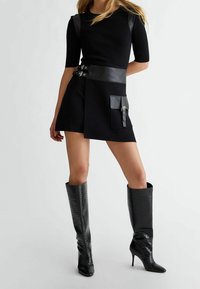 Černý pletený top s krátkými rukávy, doplněný černou sukní s koženým páskem a kapsou, doplněný kozačkami ke kolen�ům s krokodýlím vzorem.