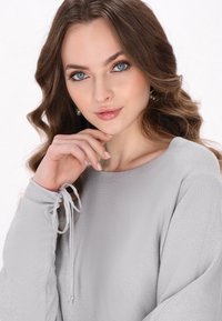Maglietta grigia a maniche lunghe con una texture morbida, caratterizzata da dettagli annodati ai polsini e un collo arrotondato. Il materiale ha una leggera lucentezza.