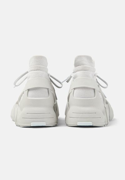 Paire de baskets blanches montantes avec des chevilles type chaussettes et des lacets, vues de dos sur un fond blanc.