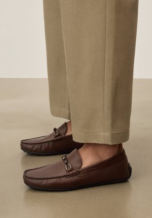 Mocassins en cuir marron avec détail en métal, portés avec un pantalon beige raccourci sur une surface de sol lisse.