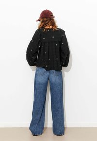 Haut noir à manches longues brodé avec un dos froncé ; associé à un jean bleu à jambes larges. Casquette bordeaux sur la tête. Fond blanc uni.