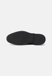 Semelle de chaussure habillée noire avec grip texturé, bout arrondi et talon, affichant la marque "Tommy Hilfiger" au centre.