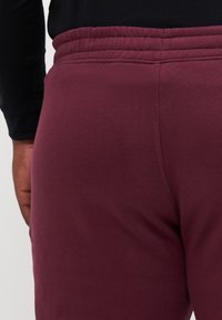 Pantalones de chándal burdeos con una textura suave, que cuentan con una cintura elástica y sutiles detalles de costura, visibles parcialmente desde la vista lateral.