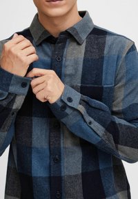 Camisa de franela en cuadros de azul, gris y azul marino. Presenta un cuello abotonado, un bolsillo frontal y puños abotonados con botones oscuros.
