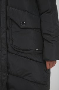 Sort pustjakke med quiltet tekstur, der har en stor sidelomme med øje og afrundede manchetter. Stoffet er glat og skinnende.