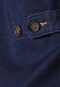 Marineblauer Stoff, der eine Nahaufnahme von zwei braunen Knöpfen mit goldener Verzierungen zeigt, die eine strukturierte Oberfläche und einen genähten Akzent aus goldenem Faden aufweisen.