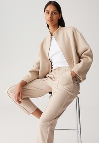 Femme assise sur un tabouret en métal, portant une veste zippée beige, un haut blanc et un pantalon beige, avec une main dans la poche et l'autre reposant sur le genou.