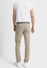 Beige Chinos mit slim fit und Gesäßtaschen, getragen mit einem weißen Hemd. Ausgestattet mit einem schwarzen Gürtel und weißen Sneakers. Glatte, Baumwollstoffe.