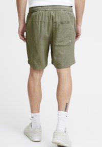 Shorts verts texturés avec une taille élastique et une poche arrière, présentant une coupe décontractée et un ourlet roulé. Le matériau semble léger et respirant.