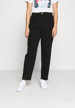 Vrouw draagt een high-waisted zwarte jeans met rechte pijpen, witte sneakers en een wit T-shirt met de tekst "Total Catch".