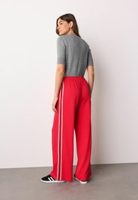 Haut gris à manches courtes avec un détail texturé associé à un pantalon large rouge doté de bandes latérales blanches. Le mannequin se tient de côté sur une surface neutre.