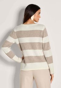 Frau mit langen dunklen Haaren, die einen beigen und weißen gestreiften Pullover und helle beige Hosen trägt, steht mit einer Hand auf der Hüfte und blickt weg.