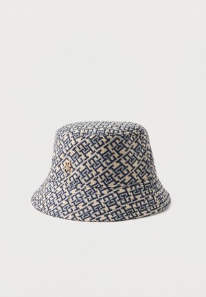 Sombrero bucket con patrón geométrico azul y beige y pequeño emblema dorado en el lado, mostrado sobre un fondo blanco liso.