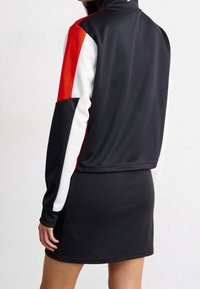Svart atletisk jacka med röda och vita ärmdekorer, hög krage, figurnära passform, kombinerad med en svart kjol. Slät tygtextur.