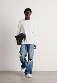 Hvit langermet skjorte med lite logo, slitte blåjeans med store rifter, holder en svart clutch veske, kombinert med hvite joggesko.