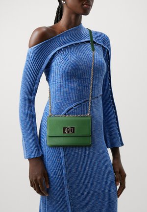 Sac bandoulière en cuir vert avec une chaîne et quincaillerie noire. Surface texturée et forme rectangulaire. Le modèle porte une robe bleue à côtes.