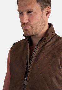 Brown leather vest with quilted design, high collar, and silver zipper detail, layered over a maroon sweater. Smooth texture.