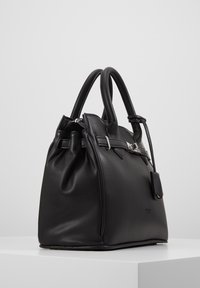 Sac à main en cuir noir avec deux poignées, détail de boucle argentée et petite étiquette attachée, posé sur une surface blanche sur un fond uni.