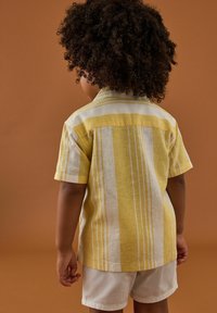 Camisa de manga corta a rayas amarillas y blancas, hecha de tejido ligero. Presenta un cuello con botón y un yugo en la parte posterior. Combinada con pantalones cortos blancos.