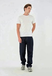 T-shirt branca de mangas curtas combinada com jeans largos de cor azul escura e ténis cinzentos, de pé contra um fundo simples.