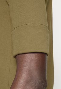 Vihreä puuvillainen pitkähiha paita, jossa on käännetyt hihansuut. Kangas on pehmeä, sileä ja sopii tiiviisti ranteen ympärille, korostaen siistiä saumaa.