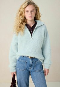 Junge Frau mit langen, lockigen blonden Haaren, die einen hellblauen, flauschigen Reißverschluss-Pullover über einem dunklen Spitzenoberteil trägt, dazu blaue Jeans und eine kastanienbraune Tasche hält.