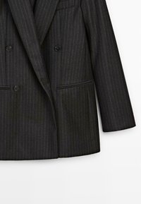 Blazer a righe nere con design doppiopetto, due tasche frontali e bottoni neri. Tessuto liscio e strutturato.