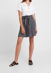 Femme portant une chemise blanche à manches courtes boutonnée, une jupe grise mi-genou avec ceinture nouée, et des sandales noires à enfiler, debout sur fond blanc.
