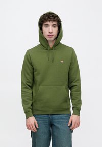 Πράσινο βαμβακερό φούτερ με κουκούλα και τσέπη στο μπροστινό μέρος, κορδόνι στην κουκούλα και μικρό λογότυπο. Ομαλή υφή και άνετη εφαρμογή. Φορεμένο με μπλε τζιν.