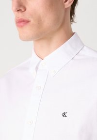 Λευκό βαμβακερό πουκάμισο με κουμπιά και κλασικό γιακά, διαθέτοντας ένα μικρό μαύρο λογότυπο στην αριστερή πλευρά του στήθους. Λείο ύφασμα και εφαρμοστό κόψιμο.