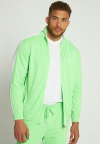 Grøn træningsdragt lavet af strækbart materiale, der inkluderer en jakke med lynlås og hvide detaljer samt matchende bukser, med en hvid T-shirt under.