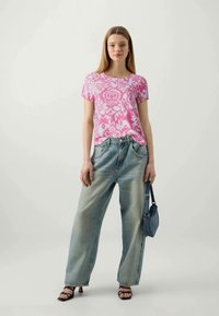 T-shirt a fiori rosa, vestibilità rilassata, abbinato a jeans a gamba larga blu chiaro. Il modello porta una piccola borsa blu e indossa sandali neri.