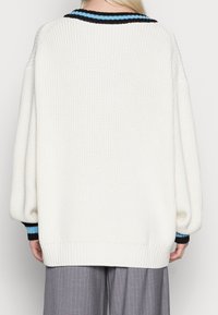 Pull en tricot blanc surdimensionné avec des poignets côtelés, présentant un col rayé noir et bleu. Texture lisse et coupe décontractée.