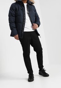 Marineblå puffjakke med aftagelig pelsforet hætte, lynlås lukning og elastiske manchetter, kombineret med sorte bukser og sorte sneakers.
