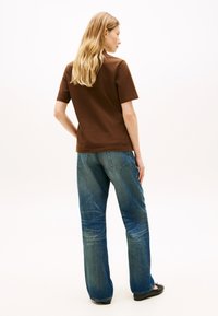 Γυναίκα με πλάτη, φορώντας καφέ  κοντομάνικο πουκάμισο, μπλε τζιν με πίσω τσέπες και μαύρα επίπεδα παπούτσια σε λευκό φόντο.