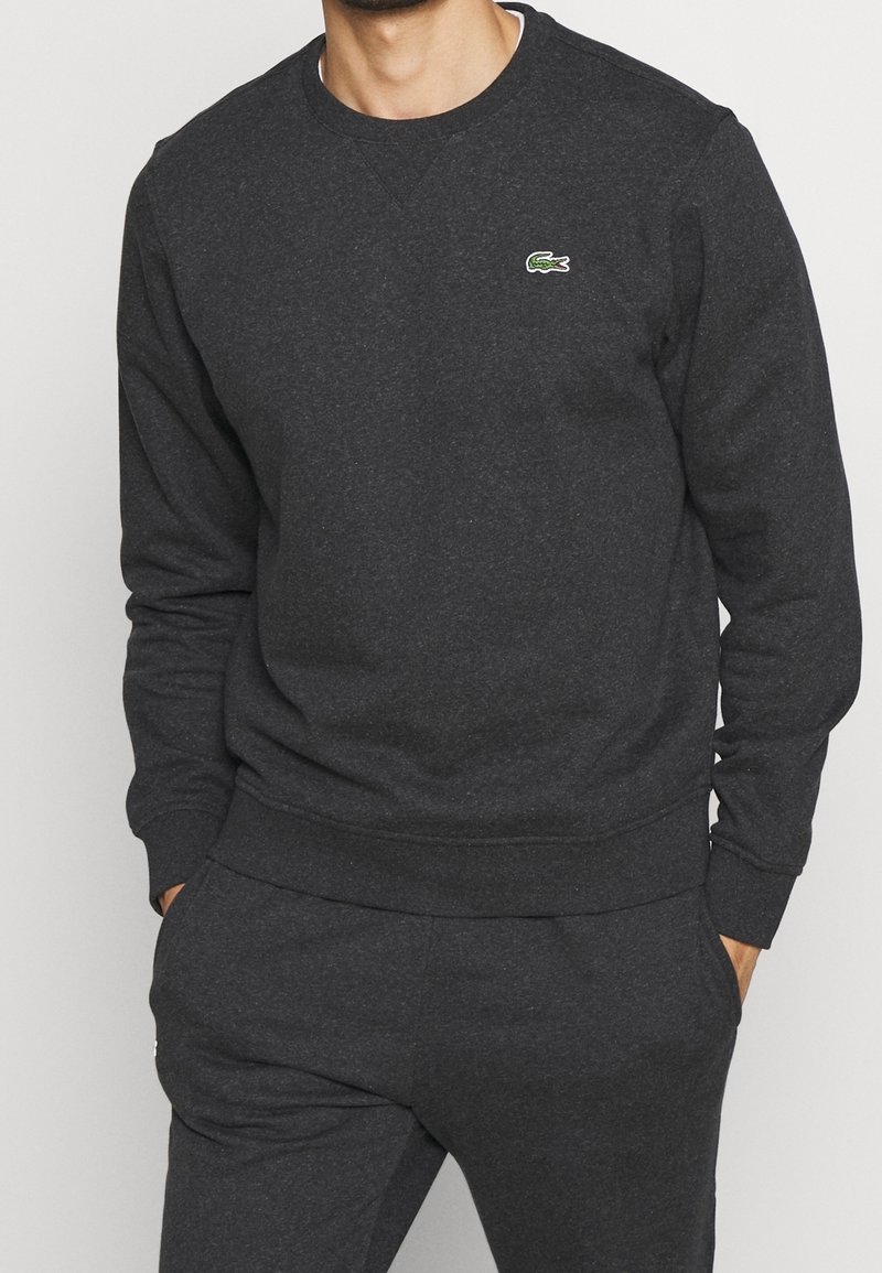 Uomo che indossa un felpa grigio scuro e pantaloni abbinati con le mani nelle tasche, caratterizzata da un piccolo logo di coccodrillo verde sul petto.