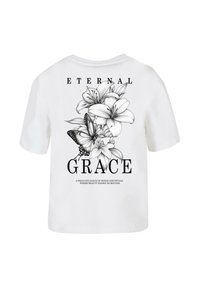Hvid bomulds T-shirt med et sort blomsterdesign med liljer og en sommerfugl. Teksten "ETERNAL GRACE" står under grafikken.