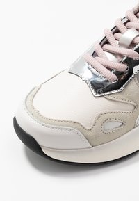 Zapatilla con un upper de cuero texturizado en beige y blanco, acentos metálicos plateados, y cordones rosa claro. Suela de goma negra.
