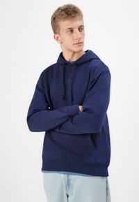 Felpa blu navy realizzata in tessuto morbido, con tasca frontale, cappuccio regolabile con cordino e logo tono su tono sul petto. Vestibilità casual.
