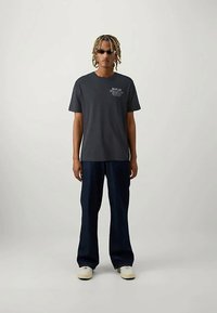 Šedé bavlněné tričko s logem "REPLAY", spárované s volnými tmavě modrými džíny a bílými teniskami. Model má na sobě sluneční brýle.