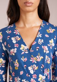 Blå omlottklänning med blommönster i rosa, gult och vitt. Har en djup V-halsringning och långa ärmar, tillverkad av lätt tyg.