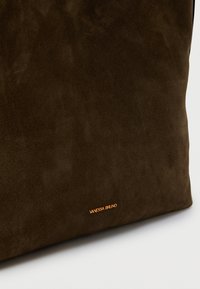 Τσάντα χειρός από σκούρο καφέ σουέντ με μικρό χρυσό λογότυπο «Vanessa Bruno» κοντά στο κάτω άκρο, τοποθετημένη πάνω σε απλό ανοιχτόχρωμο φόντο.